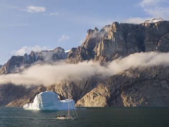 Aerial view of a sailing boat navigating along the fjords with iceberg near the coast in Sermersooq, Greenland. - AAEF17062