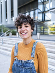 Ethnische Frau in Jeans-Overall mit zahnigem Lächeln schaut in die Kamera, während sie vor einem modernen Gebäude steht - ADSF41487