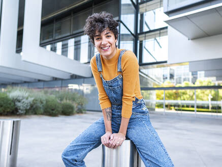 Hispanic Frau in Jeans Overall Blick auf die Kamera genüsslich beim Sitzen auf der Straße vor modernen Gebäude in der Tageszeit - ADSF41490