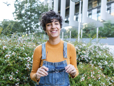 Glückliche hispanische Frau, die ihren Jeans-Overall ausbreitet und in die Kamera schaut, in der Nähe eines modernen Gebäudes in blühenden Büschen im Sommer - ADSF41493