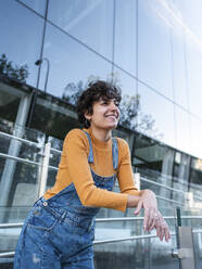 Low angle of cheerful young woman with curly short hair in denim overall standing on balcony and leaning on glass fence while looking away near urban building - ADSF41495