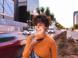 Smiling young female with curly short hair in casual clothes strolling along roadway with cars and recording voice message via cellphone during evening time - ADSF41499