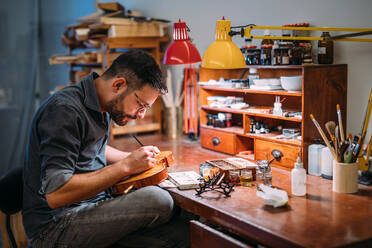 Seitenansicht eines erwachsenen Handwerkers mit Brille, der in einer Geigenbauwerkstatt am Tisch sitzt und im Lampenlicht eine zusammengebaute Holzgeige färbt - ADSF41502