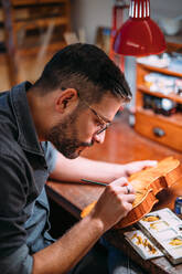 Seitenansicht eines erwachsenen Handwerkers mit Brille, der in einer Geigenbauwerkstatt am Tisch sitzt und im Lampenlicht eine zusammengebaute Holzgeige färbt - ADSF41503