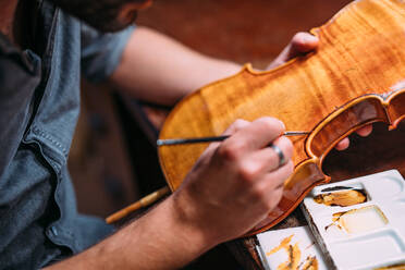 Cropped unrecognizable adult craftsman sitting at table in luthiery and coloring assembled wooden violin in lamplight - ADSF41504