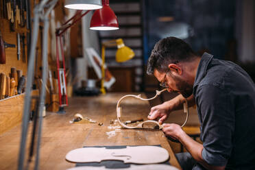 Seitenansicht eines konzentrierten Handwerkers mit Brille, der an der Werkbank sitzt und eine neue Geige baut, beginnend mit der Formung der Rippen - ADSF41505