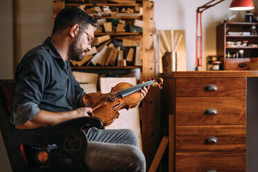 Seitenansicht eines erwachsenen Holzarbeiters mit Brille, der eine handgefertigte klassische Geige hält, während er in einem Sessel in seinem eigenen Geigenbauatelier sitzt - ADSF41508