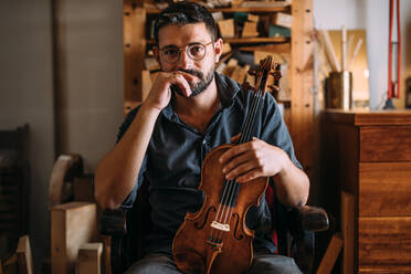 Nachdenklicher erwachsener Holzarbeiter mit Brille, der eine handgefertigte klassische Geige in der Hand hält, während er in einem Sessel im eigenen Geigenbauatelier sitzt und in die Kamera schaut - ADSF41509