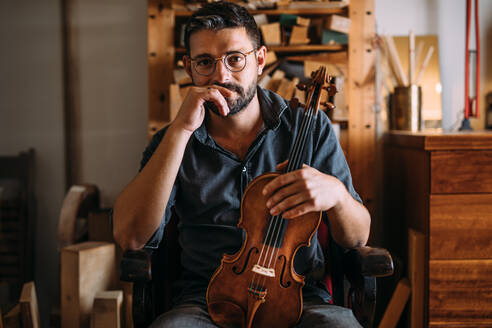 Nachdenklicher erwachsener Holzarbeiter mit Brille, der eine handgefertigte klassische Geige in der Hand hält, während er in einem Sessel im eigenen Geigenbauatelier sitzt und in die Kamera schaut - ADSF41509