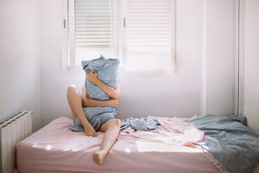 Full body of unrecognizable barefoot female sitting on comfortable bed near window and embracing pillow while covering face in morning - ADSF41513