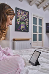 Reife Frau in rosa Pyjama sitzt auf dem Bett und liest ein Buch in sonnigen Wochenende Morgen zu Hause - ADSF41515