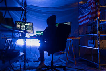 Eine Rückansicht eines vermummten Hackers am Computer in einem dunklen Raum bei Nacht, Cyberwar-Konzept. - HPIF00977