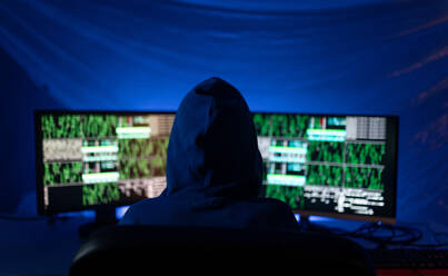 Eine Rückansicht eines vermummten Hackers am Computer in einem dunklen Raum bei Nacht, Cyberwar-Konzept. - HPIF00978