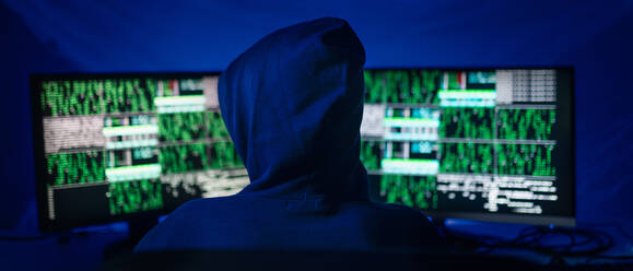 Eine Rückansicht eines vermummten Hackers am Computer in einem dunklen Raum bei Nacht, Cyberwar-Konzept. - HPIF00979