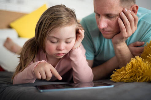 Ein Vater und seine kleine Tochter mit Down-Syndrom liegen auf dem Bett und benutzen zu Hause ein Tablet. - HPIF01019