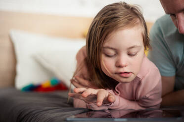 Ein Vater und seine kleine Tochter mit Down-Syndrom liegen auf dem Bett und benutzen zu Hause ein Tablet. - HPIF01020
