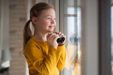 A curious little girl with Down syndrome with binoculars looking through window at home. - HPIF01044