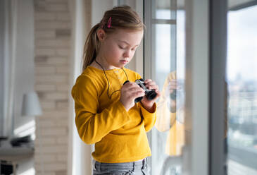 Ein neugieriges kleines Mädchen mit Down-Syndrom schaut mit einem Fernglas durch ein Fenster zu Hause. - HPIF01045