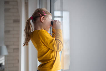 Ein neugieriges kleines Mädchen mit Down-Syndrom schaut mit einem Fernglas durch das Fenster zu Hause. - HPIF01046