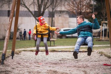 Ein Vater mit seiner kleinen Tochter mit Down-Syndrom auf einer Schaukel im Freien in einem Spielplatz. - HPIF01075