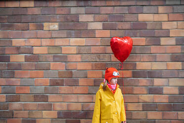 A little girl with Down syndrome holding heart shaped balloon in winter against brick wall. - HPIF01084