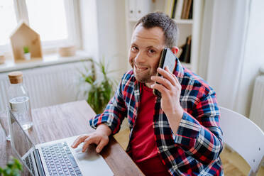 Ein junger Mann mit Down-Syndrom sitzt zu Hause am Schreibtisch und benutzt Smartphone und Laptop, schaut in die Kamera und lächelt. Konzept der Integration behinderter Menschen, Home Office. - HPIF01095