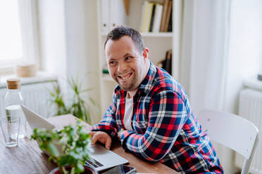 A young man with Down syndrome sitting at desk at home and using laptop, looking at camera and smiling. - HPIF01096