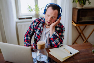 Ein junger Mann mit Down-Syndrom sitzt am Schreibtisch im Büro, benutzt einen Laptop und hört Musik über Kopfhörer. - HPIF01098