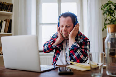 A young man with Down syndrome sitting at desk in office and using laptop, listening to music from headphones. - HPIF01099