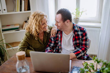 Young man with Down syndrome sitting at desk in office and using laptop, his mother assisting him. - HPIF01101