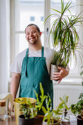 Ein junger Mann mit Down-Syndrom, der sich zu Hause um Pflanzen kümmert, lächelt und schaut in die Kamera. - HPIF01103