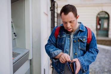 Ein glücklicher junger Mann mit Down-Syndrom benutzt einen Geldautomaten auf der Straße und hebt Geld ab. - HPIF01116