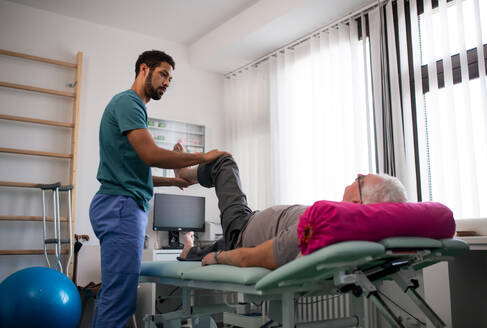 Eine Physiotherapeutin trainiert mit dem Bein eines älteren Patienten in einem Behandlungsraum. - HPIF01195