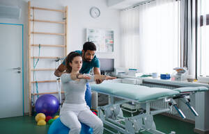 Ein junger männlicher Physiotherapeut übt mit einer jungen Patientin auf einem Ball in einem Krankenzimmer - HPIF01200