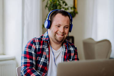 A young man with Down syndrome sitting at desk in office and using laptop, listening to music from headphones. - HPIF01203