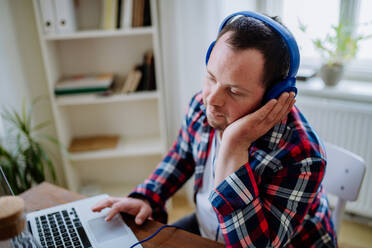 A young man with Down syndrome sitting at desk in office and using laptop, listening to music from headphones. - HPIF01204