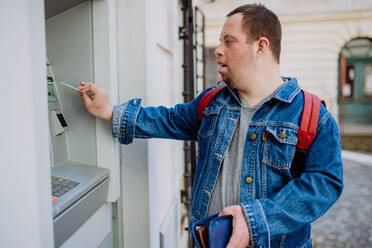 Ein glücklicher junger Mann mit Down-Syndrom benutzt einen Geldautomaten auf der Straße und hebt Geld ab. - HPIF01214