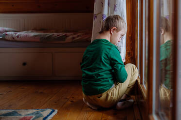 Ein kleiner Junge mit Down-Syndrom sitzt auf dem Boden und schaut durch das Fenster seines Hauses. - HPIF01244