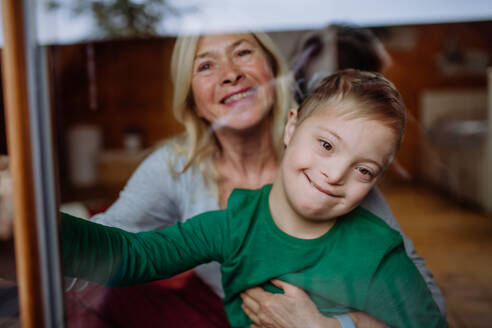 Ein Junge mit Down-Syndrom und seine Großmutter schauen durch das Fenster ihres Hauses in die Kamera. - HPIF01250