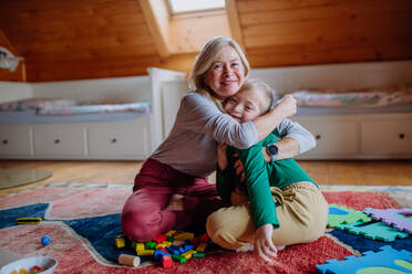 Ein glücklicher Junge mit Down-Syndrom, der auf dem Boden sitzt und sich mit seiner Großmutter zu Hause umarmt - HPIF01255