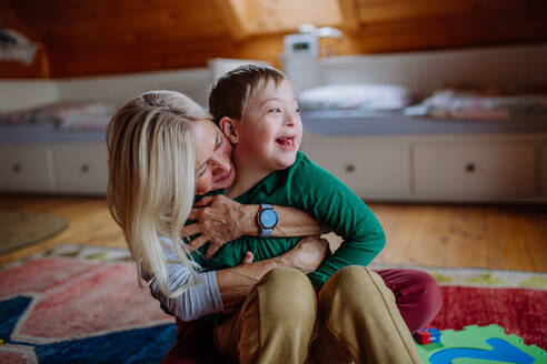 Ein glücklicher Junge mit Down-Syndrom, der auf dem Boden sitzt und sich mit seiner Großmutter zu Hause umarmt - HPIF01256