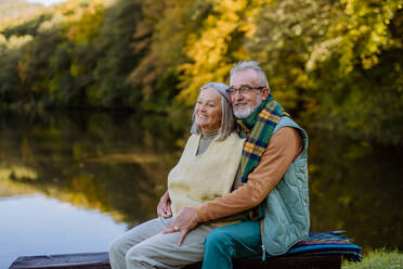 Verliebtes älteres Paar sitzt zusammen auf einer Bank in der Nähe eines Sees, während eines Herbsttages. - HPIF01331