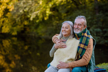 Verliebtes älteres Paar sitzt zusammen auf einer Bank in der Nähe eines Sees, während eines Herbsttages. - HPIF01333