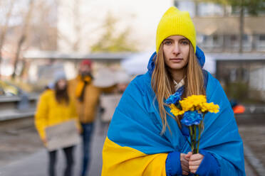 Protest against Russian invasion of Ukraine. A young woman wrapped in Ukrainian flag holding blue and yellow flowers. - HPIF01411
