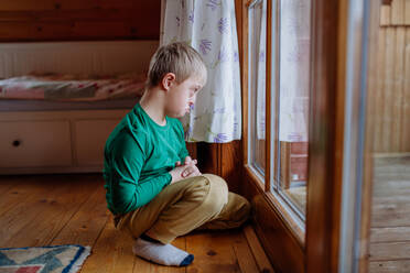 Ein trauriger kleiner Junge mit Down-Syndrom, der auf dem Boden sitzt und durch das Fenster seines Hauses schaut. - HPIF01434