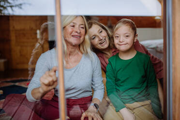 A boy with Down syndrome with his mother and grandmother sitting and looking through window at home. - HPIF01437