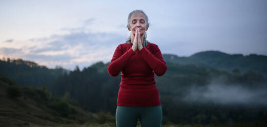 A senior woman doing breathing exercise in nature on early morning with fog and mountains in background. - HPIF01469