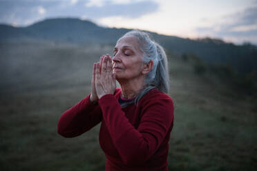 A senior woman doing breathing exercise in nature on early morning with fog and mountains in background. - HPIF01470