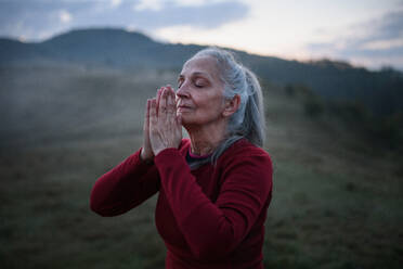Eine ältere Frau macht Atemübungen in der Natur am frühen Morgen mit Nebel und Bergen im Hintergrund. - HPIF01470