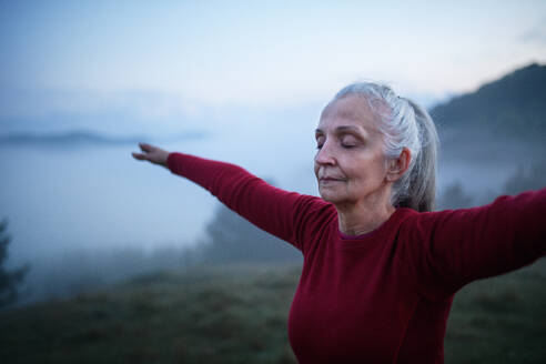 A senior woman doing breathing exercise in nature on early morning with fog and mountains in background. - HPIF01473